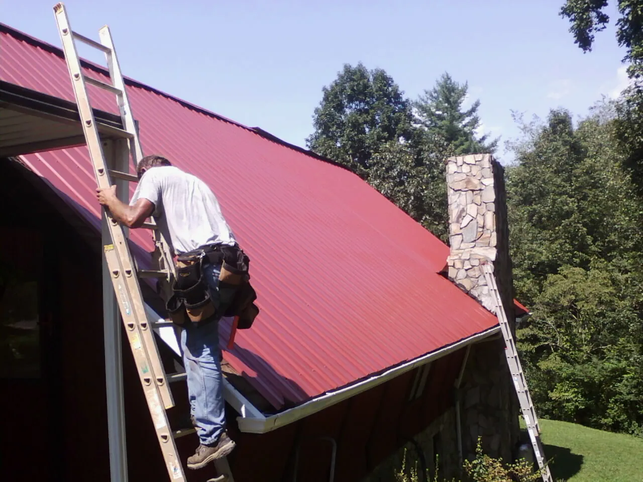 Roofer on a ladder beside a red metal roof and stone chimney — Soffit & Fascia Repair by Roofing Brothers in Cumberland Center